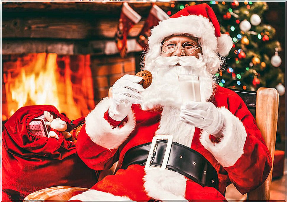 Santa Claus eating cookies and milk in front of a fireplace.